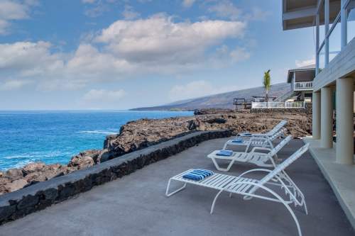 Lounge chairs on a patio overlooking rocky coastline and blue ocean under a partly cloudy sky.