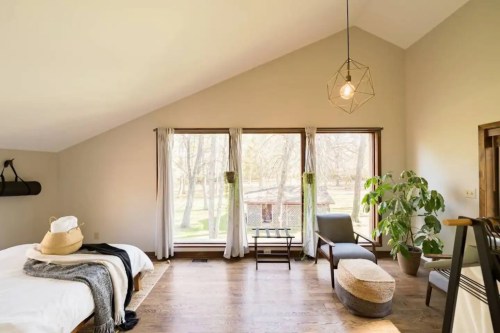 Bright, airy bedroom with large windows, a cozy bed, a chair, and a potted plant, set in a serene, natural environment.