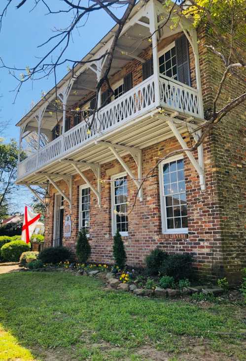 Historic brick building with a white balcony, surrounded by greenery and flowers, featuring an English flag.