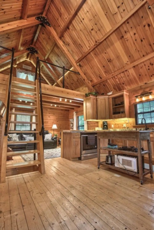 Cozy wooden cabin interior featuring a kitchen, dining area, and staircase leading to a loft. Warm, inviting atmosphere.
