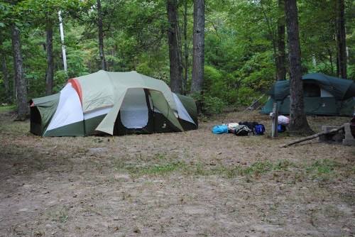 Two tents set up in a wooded campsite, surrounded by trees and scattered camping gear on the ground.