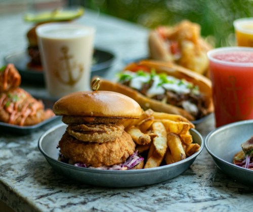A crispy chicken sandwich with onion rings, served with fries, alongside various drinks and dishes on a table.