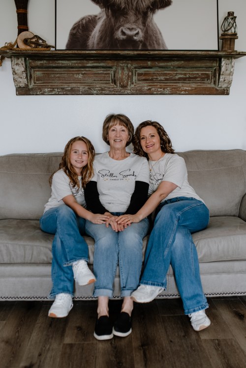 A woman sits on a couch with her two daughters, all wearing matching shirts and smiling together.