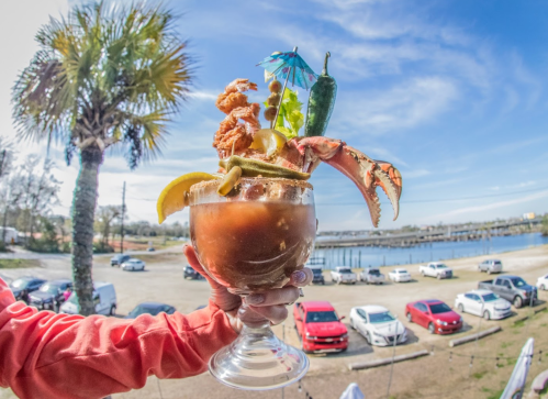 A colorful seafood cocktail garnished with shrimp, crab, and an umbrella, held against a sunny waterfront backdrop.