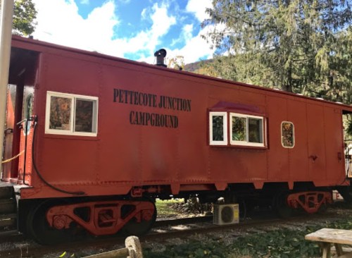 A red train caboose with windows, labeled "Pettecote Junction Campground," surrounded by trees and blue sky.