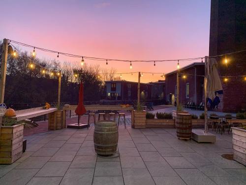 Outdoor patio at sunset, featuring string lights, wooden barrels, and a serene atmosphere with buildings in the background.