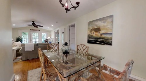 A bright dining area with a glass table, wicker chairs, and a coastal painting, leading to a cozy living room.