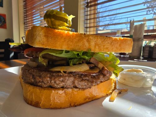 A thick burger with lettuce, tomato, pickles, and grilled onions, served on toasted bread with a side of sauce.