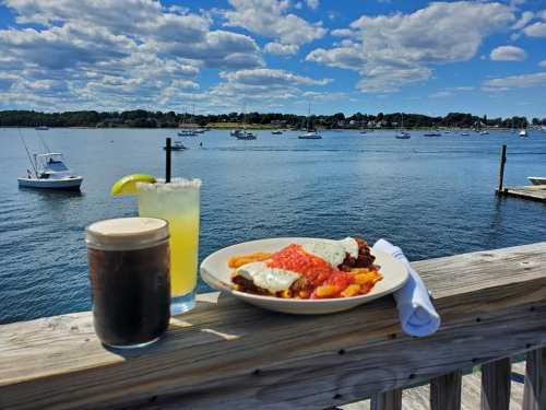 A scenic waterfront view with a plate of food and drinks on a wooden table, boats in the background under a blue sky.