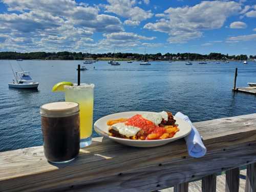 A scenic waterfront view with a plate of food and drinks on a wooden table, boats in the background under a blue sky.