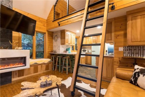 Cozy wooden cabin interior with a ladder, kitchen area, and a warm fireplace, featuring rustic decor and a cowhide rug.
