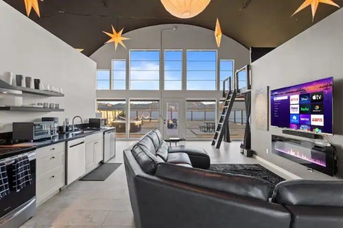 Modern living space with a black sectional sofa, large windows, and a sleek kitchen, overlooking a scenic view.