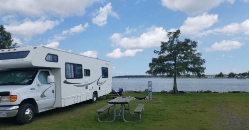 An RV parked by a lake with a picnic table and a tree under a partly cloudy sky.