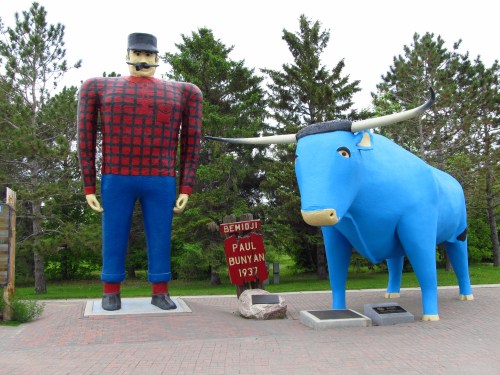 A large statue of Paul Bunyan in a plaid shirt stands next to a blue ox, with trees and a sign in the background.