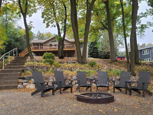 A cozy backyard with a fire pit surrounded by Adirondack chairs, leading up to a house among trees in autumn.