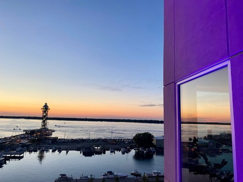 Sunset over a river with a lighthouse and boats docked, reflecting in a nearby window.