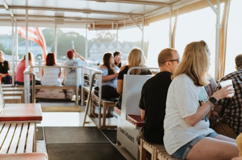People sitting on a boat, enjoying drinks and the view, with sunlight streaming in and flags visible in the background.