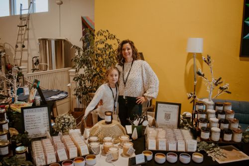 A woman and a girl stand behind a table filled with candles and jars, showcasing products in a bright, colorful space.