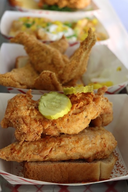 A close-up of fried chicken pieces on bread, garnished with pickles, served in a paper tray.
