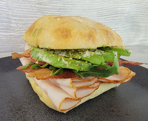 A sandwich with layers of turkey, bacon, avocado, and greens on a ciabatta roll, served on a black plate.