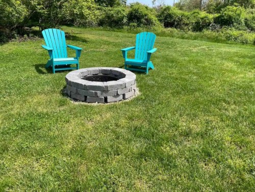 Two turquoise adirondack chairs beside a stone fire pit on a grassy lawn, surrounded by greenery.