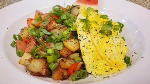 A plate of fluffy scrambled eggs with green onions, served with seasoned potatoes and a slice of watermelon.