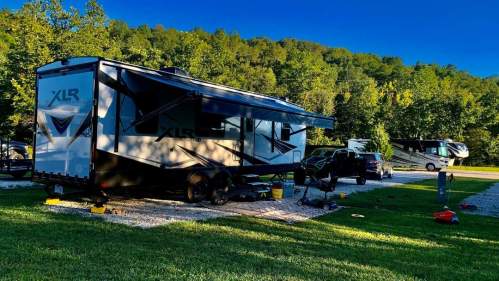 A parked RV with an awning extended, surrounded by trees and a grassy area, with tools and equipment nearby.