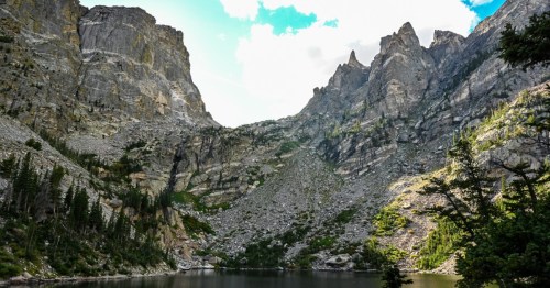 A serene mountain landscape featuring steep cliffs, a calm lake, and lush greenery under a partly cloudy sky.