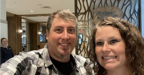A smiling couple poses for a selfie in a modern restaurant with a wooden decor background.