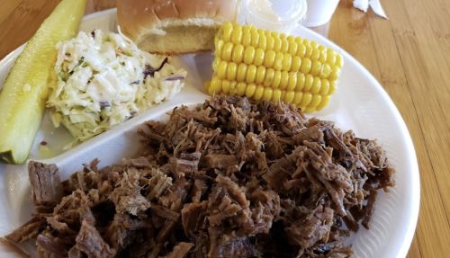A plate with shredded beef, a bun, corn on the cob, coleslaw, and a pickle slice.