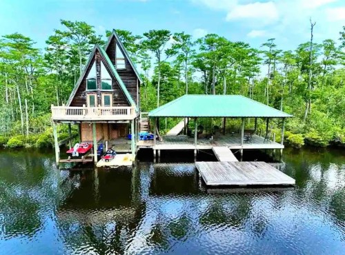 Aerial view of a unique A-frame cabin on stilts by a calm waterway, surrounded by lush green trees.