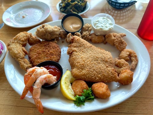 A plate of fried seafood including shrimp, fish, hushpuppies, and dipping sauces, garnished with lemon and parsley.