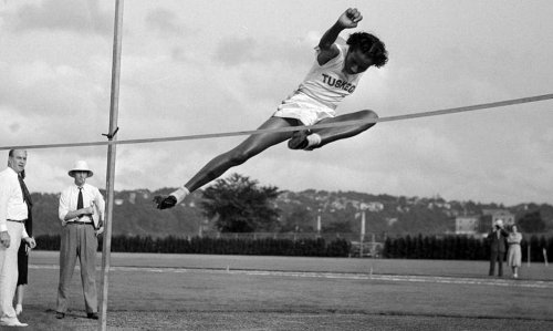 A high jumper leaps over a bar during a competition, with spectators watching in the background.