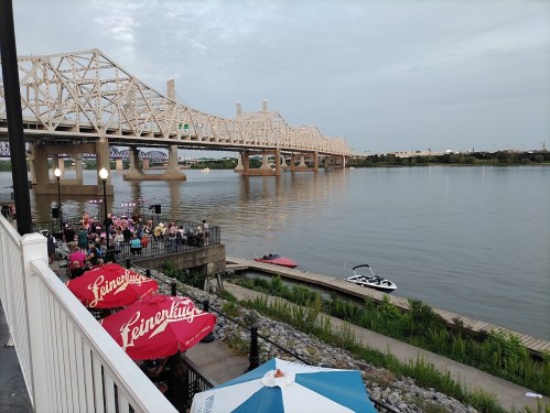 View of a river with a bridge in the background, outdoor seating with umbrellas, and people gathered by the water.