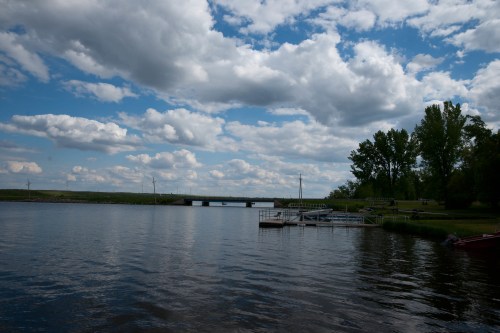 A serene river scene with a bridge in the distance, surrounded by lush greenery and a partly cloudy sky.