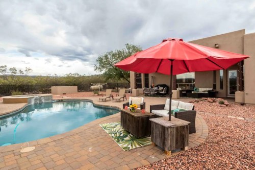 A backyard pool area with a red umbrella, seating, and a stone patio, surrounded by desert landscaping and cloudy skies.
