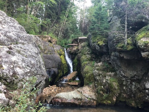 A serene waterfall cascades between moss-covered rocks in a lush, green forest.