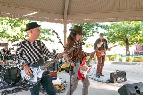 A band performs on stage under a gazebo, featuring guitarists and a bassist, with trees in the background.