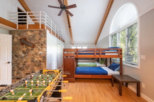 A cozy room with a bunk bed, foosball table, and large windows, featuring wooden beams and a stone wall.