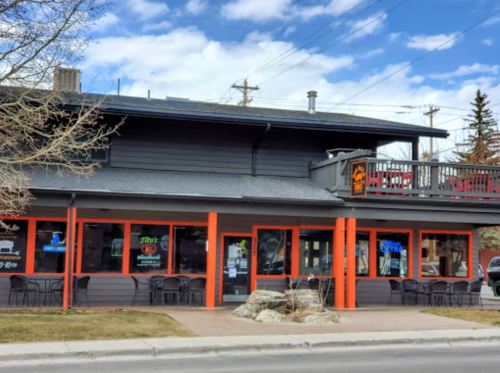A two-story building with orange trim, large windows, and outdoor seating, set against a blue sky with clouds.