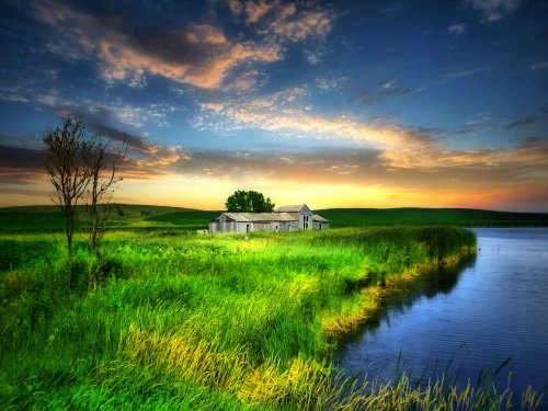 A serene landscape featuring a weathered house by a calm lake, surrounded by lush green fields and a colorful sunset sky.