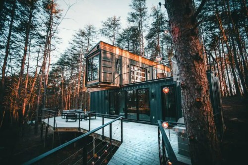 A modern, elevated cabin made from shipping containers, surrounded by trees and featuring a deck with outdoor seating.