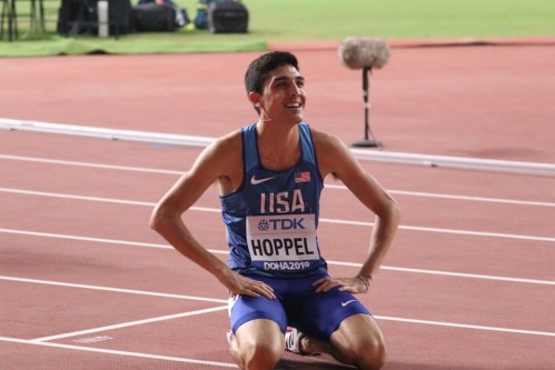A smiling athlete in a blue USA uniform sits on the track, celebrating after a race.