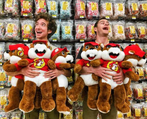 Two smiling individuals hold large stuffed animals in front of a colorful candy display.