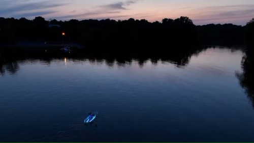 A lone kayaker paddles on a calm river at dusk, surrounded by trees and a colorful sky reflecting on the water.