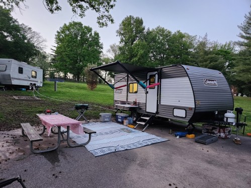 A camping trailer with an awning, picnic table, and outdoor setup in a green park area.