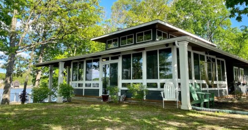 A charming lakeside cabin with large windows, surrounded by greenery and a clear blue sky.