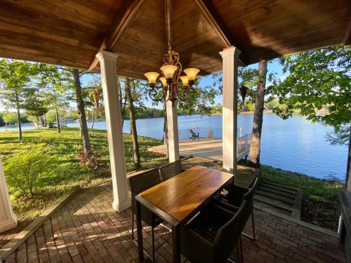 A serene lakeside view from a covered porch with a dining table and chairs, surrounded by trees and sunlight.