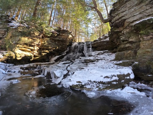 A serene waterfall cascades over rocky cliffs, surrounded by snow and lush green trees in a tranquil forest setting.