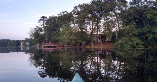 A calm lake scene with trees and houses reflected in the water, viewed from a kayak.